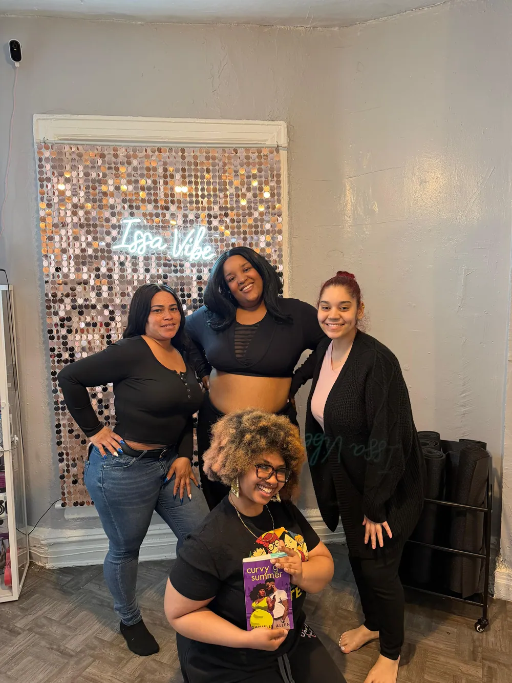 Four book club members pose in front of the studio's rose-gold sequin wall and Issa Vibe neon sign, one kneeling in front with a copy of Curvy Girl Summer