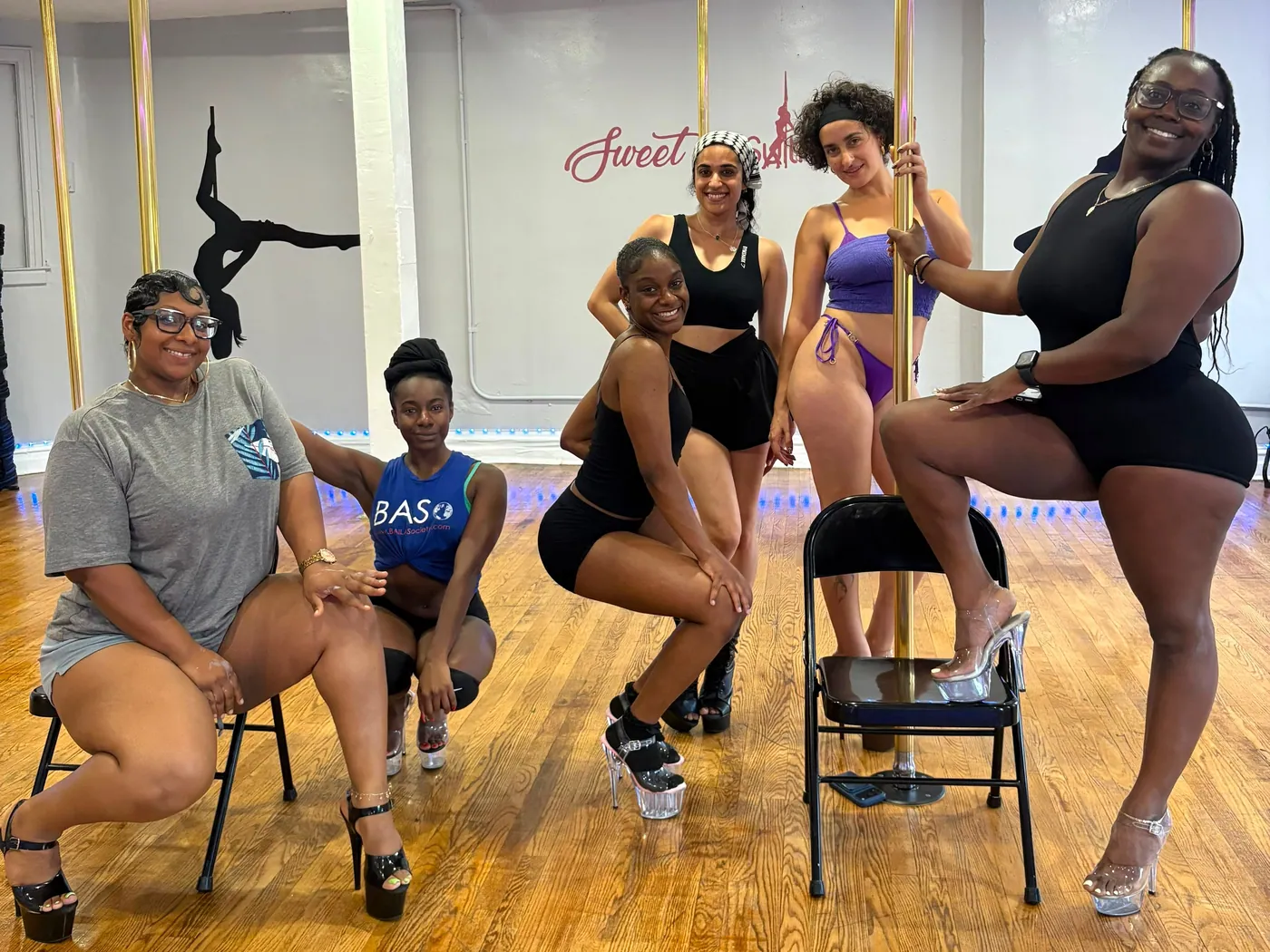 Six students in platform heels pose with folding chairs around a gold pole, one with a foot up on a chair seat