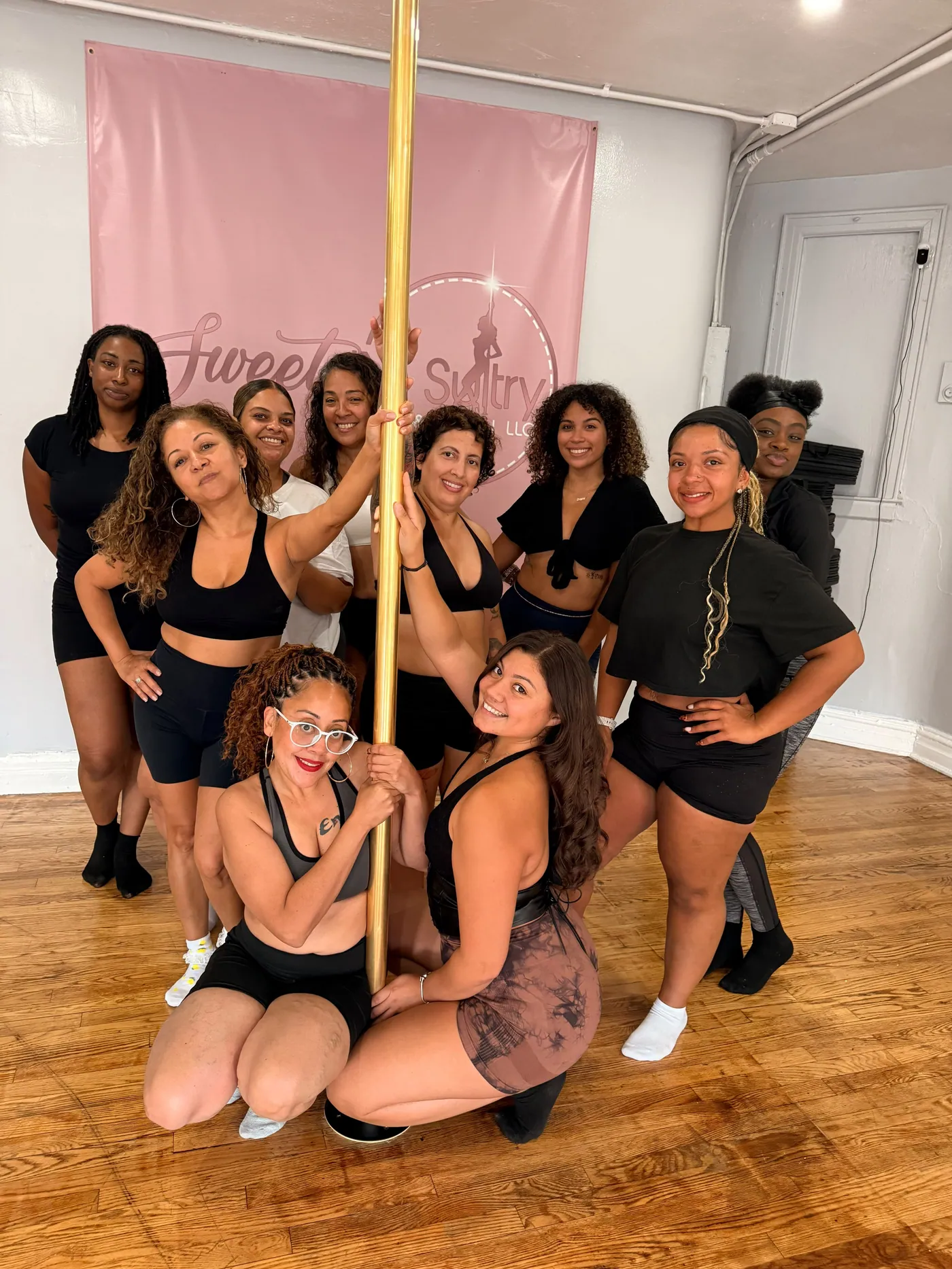 Ten students in black workout wear crowd around a gold pole in front of the studio's pink Sweet 'N Sultry banner, two crouching at the front