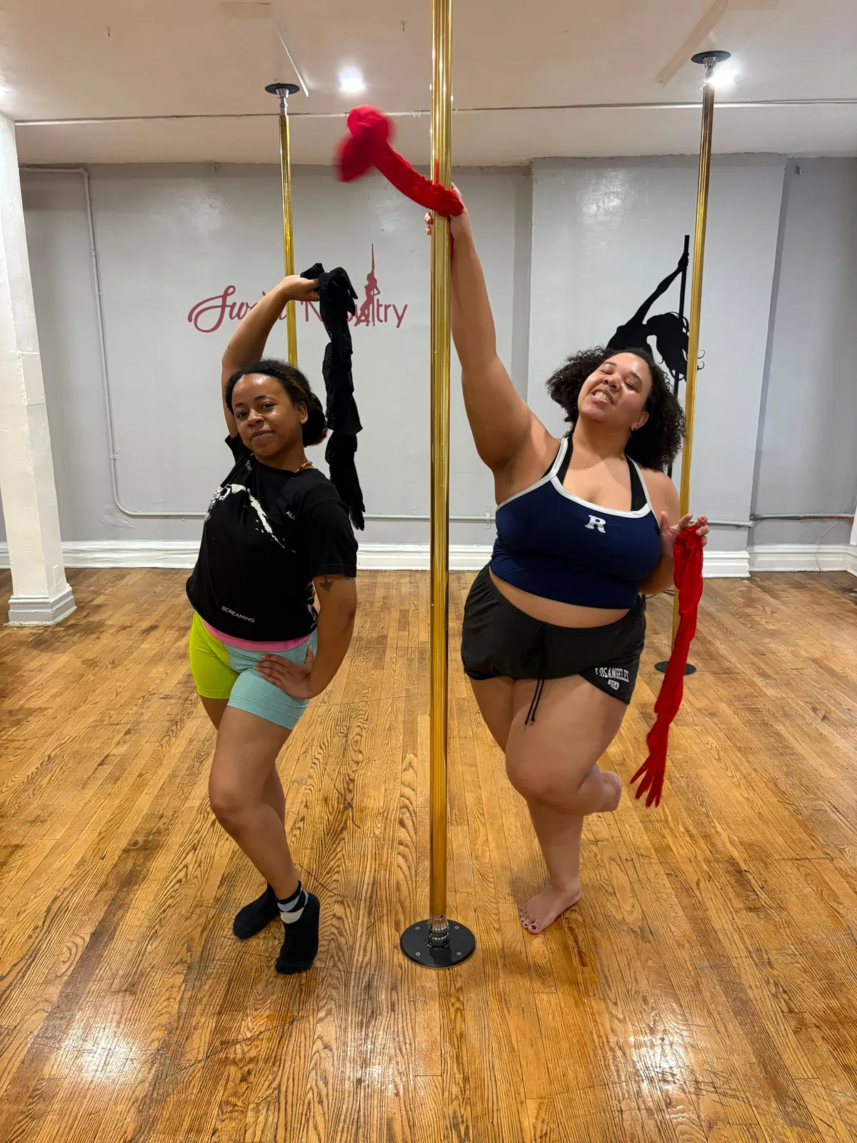 Two students twirl long burlesque gloves at a gold pole, one with a knee lifted