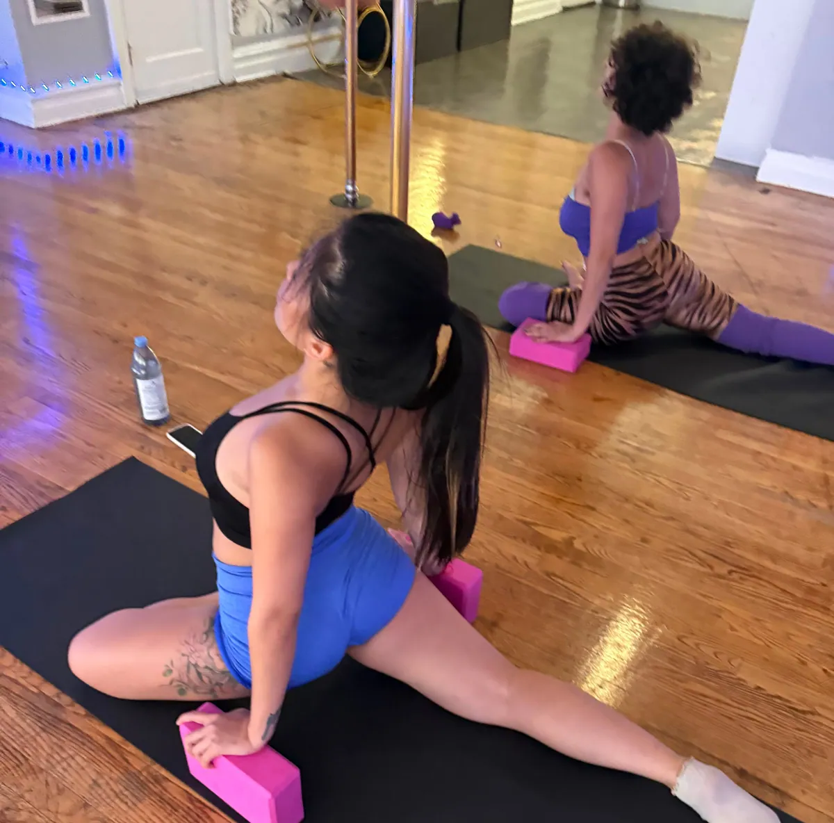 A student stretches into a half split on a mat with pink yoga blocks while another stretches on the mat behind her