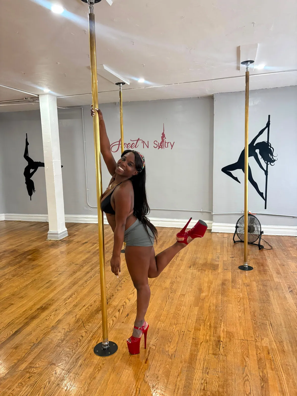 A smiling dancer in red platform heels holds a gold pole overhead and kicks one heel up behind her