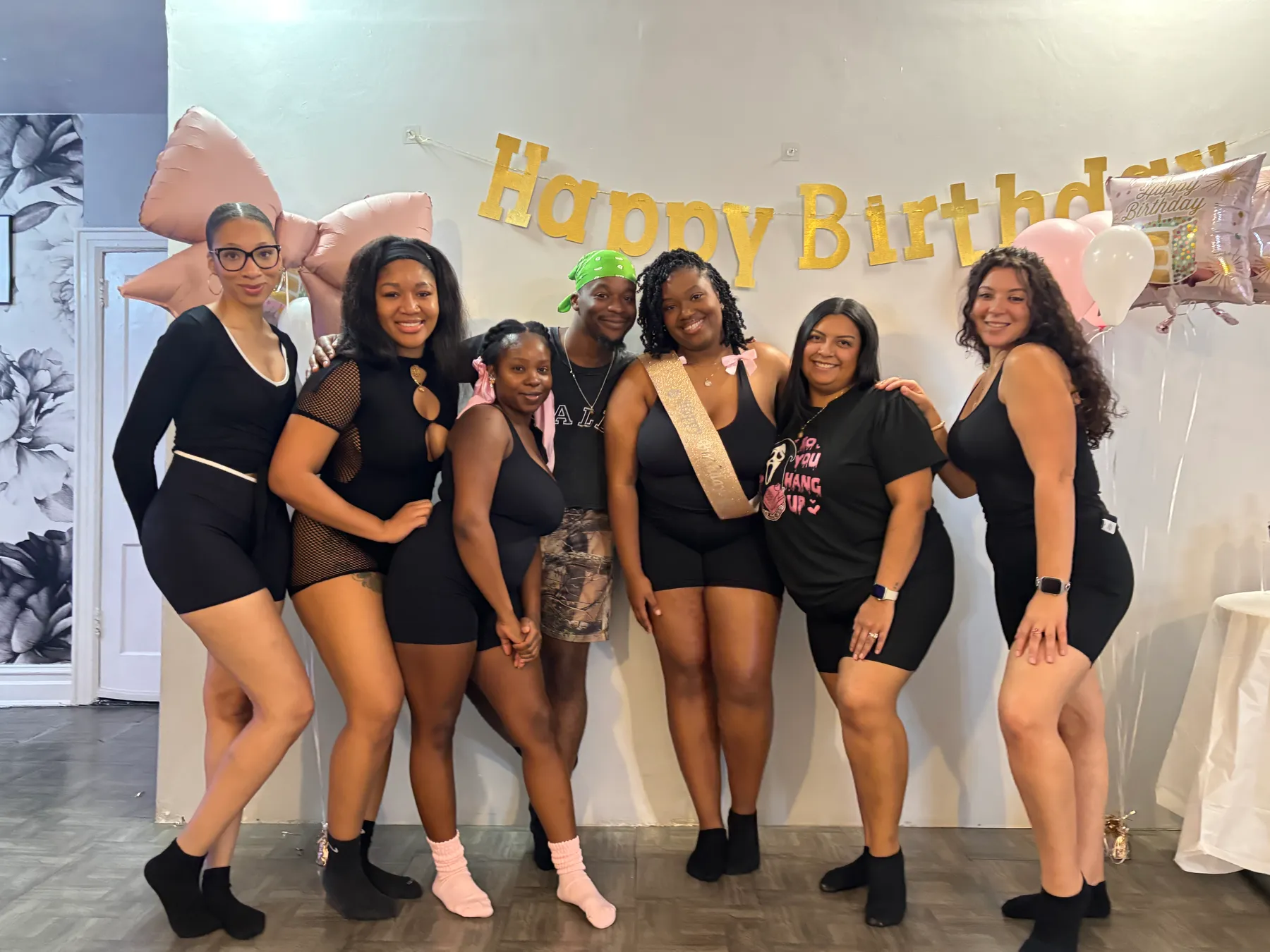 Seven friends in black pose under a gold Happy Birthday banner and a big pink bow balloon, the birthday guest wearing a gold sash