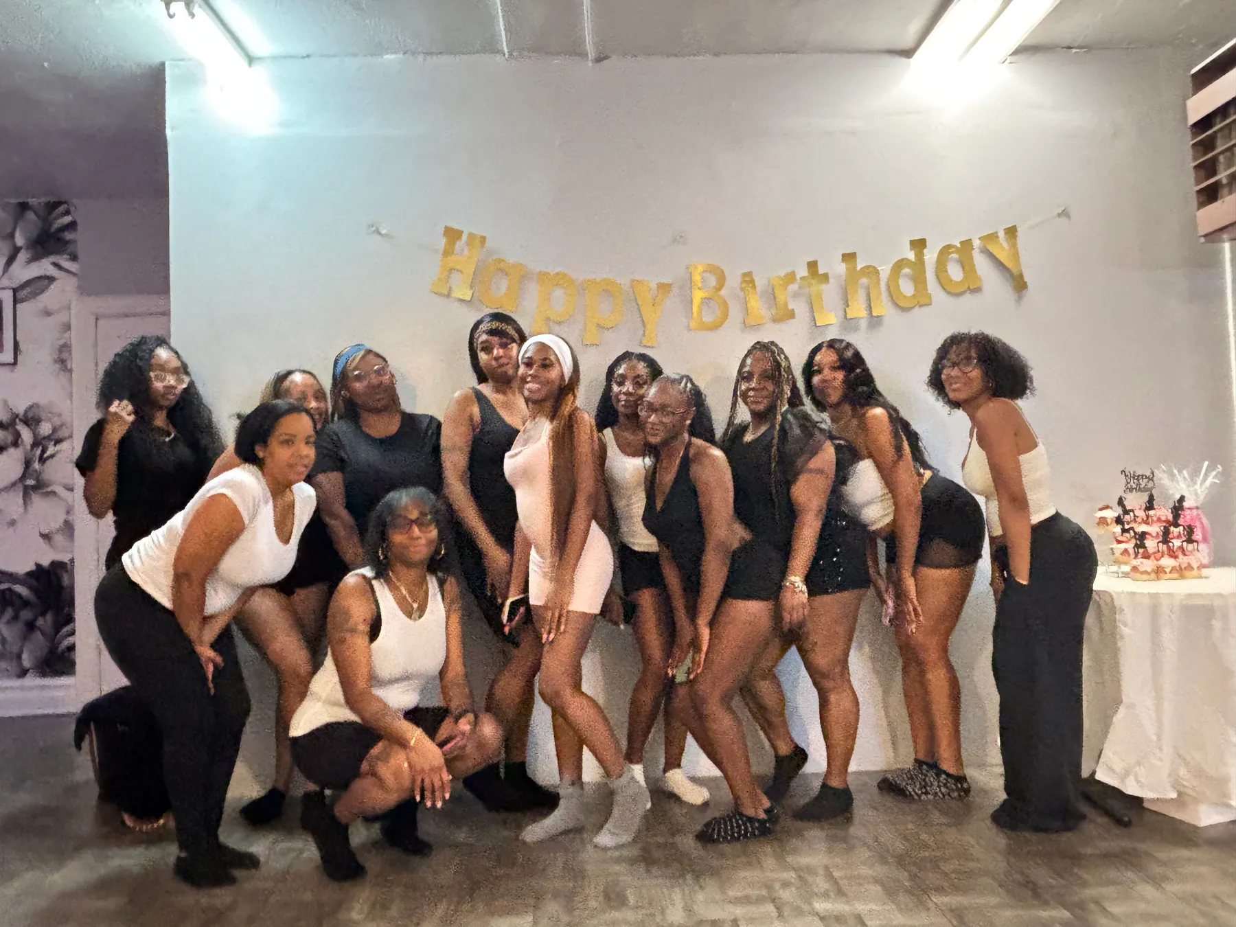 Twelve women in black and white pose under a gold Happy Birthday banner, one crouching in front, beside a cupcake stand with dancer toppers