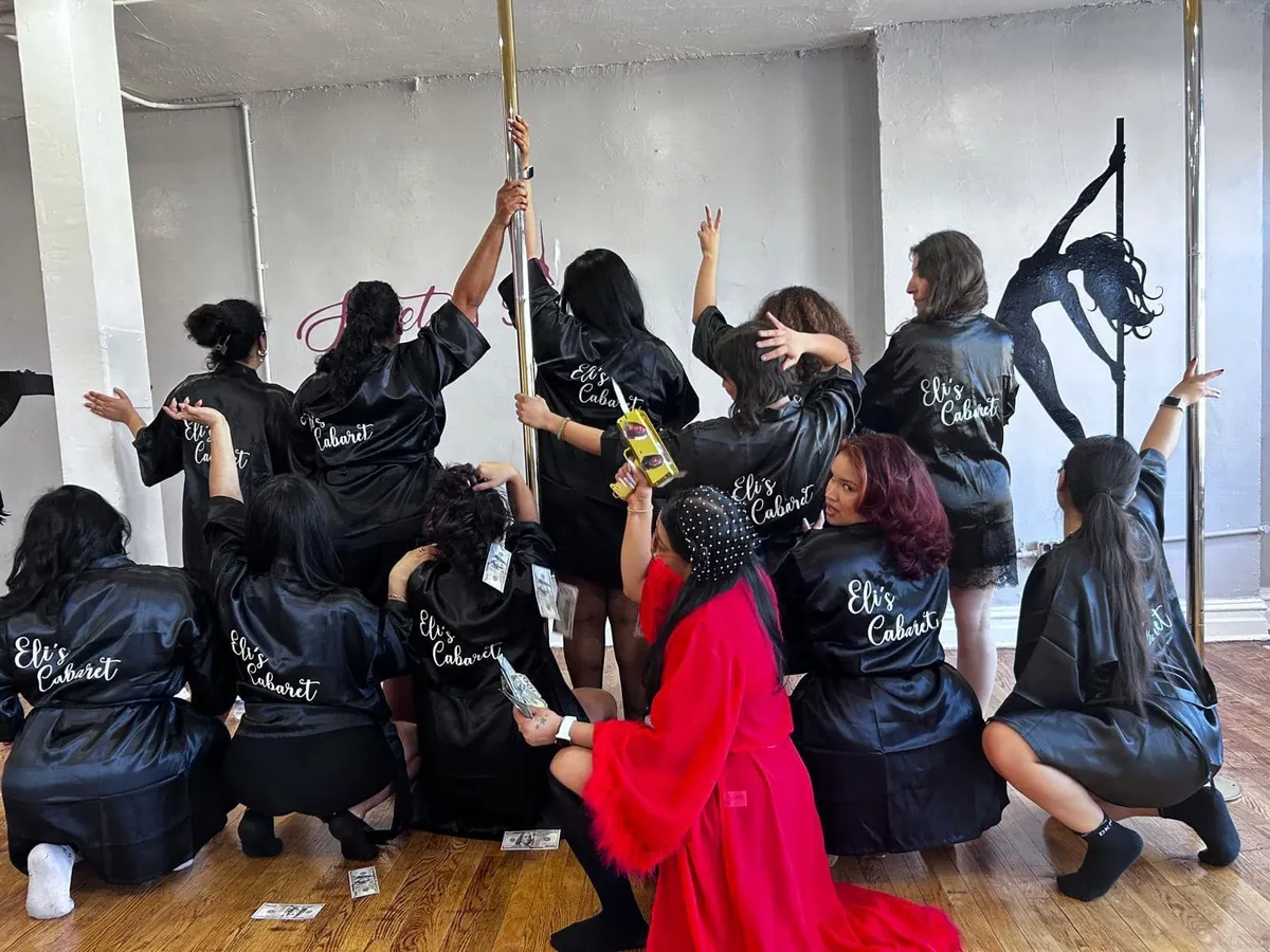Party guests in black satin robes printed “Eli's Cabaret” pose at the pole with their backs to the camera, while one in a red feather-trimmed robe kneels in front with a toy money gun held high and dollar bills on the floor