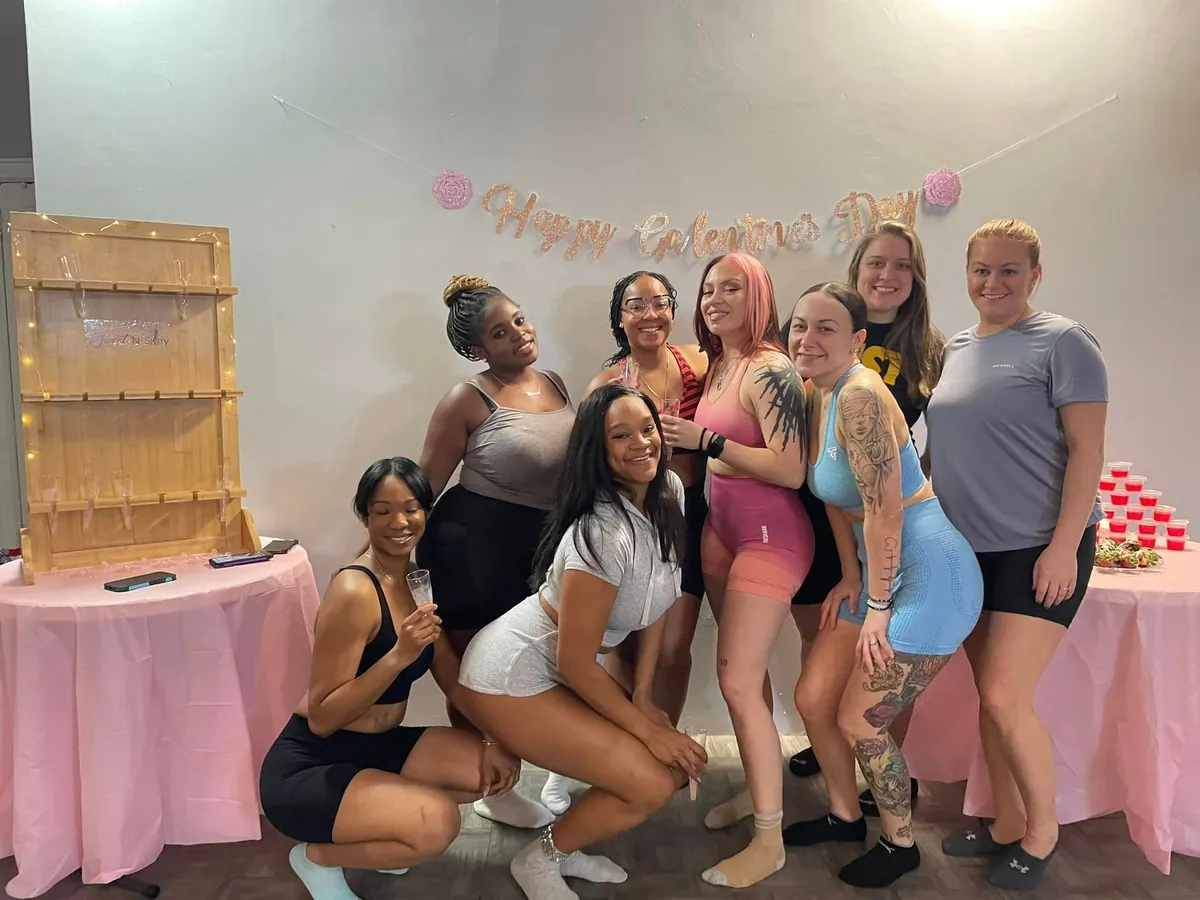 Eight friends in workout wear pose under a glittery “Happy Galentine's Day” banner, between pink-covered tables with a champagne flute wall on one and rows of red jello shots on the other