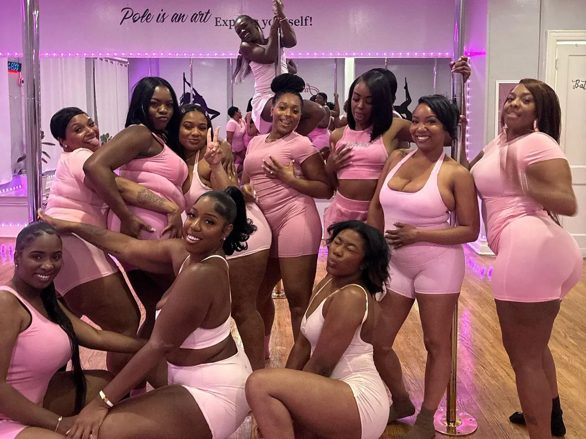 Eleven women in pink pose together under pink lights, one climbing the pole behind them, beneath a wall decal that reads “Pole is an art”